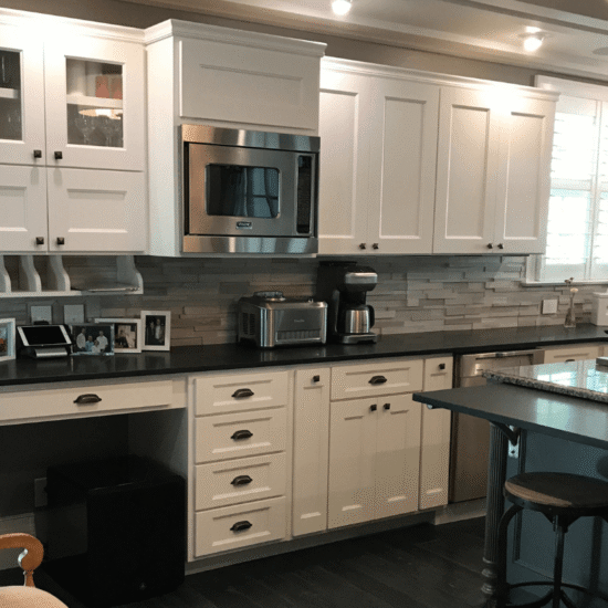 Renovated home kitchen with custom white cabinetry, stainless steel appliances, built-in desk, granite counters, coffered ceiling with accent lighting, and dark hardwood flooring.