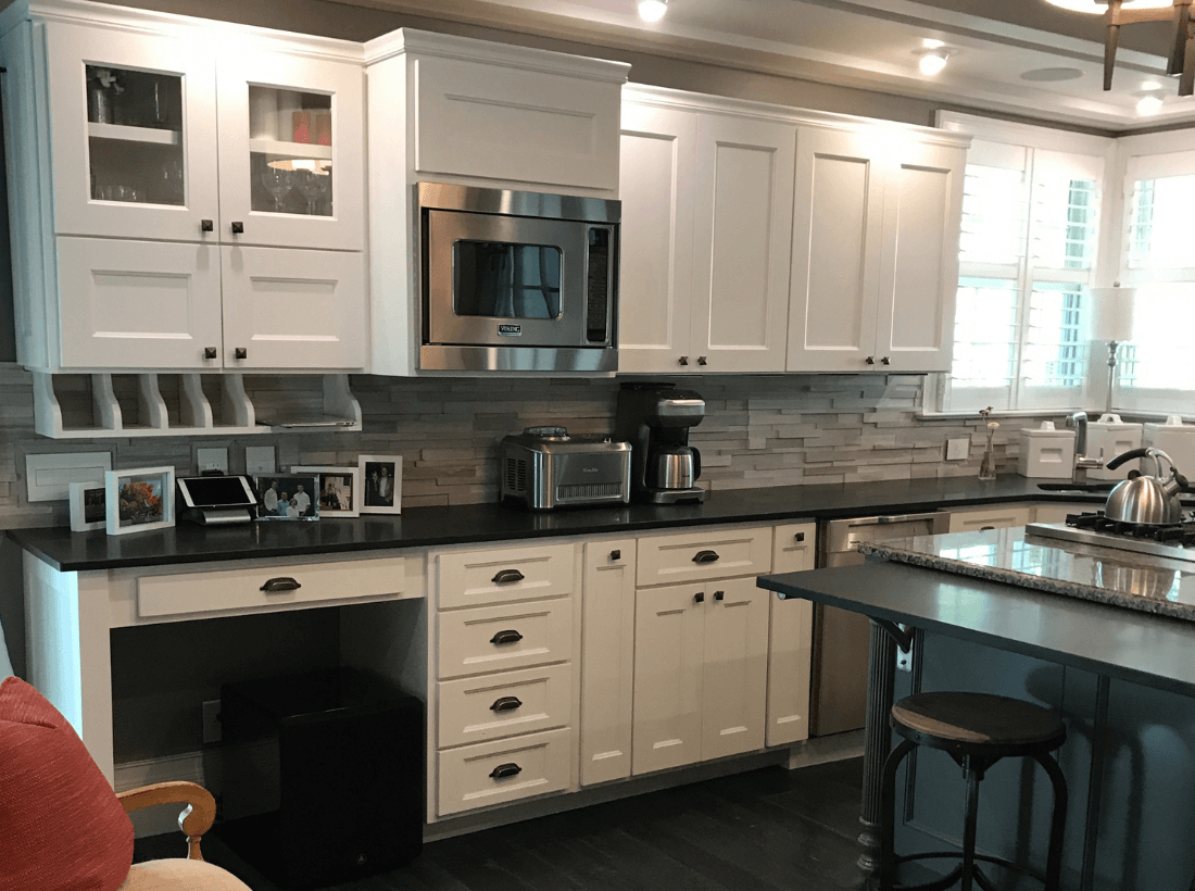 Renovated home kitchen with custom white cabinetry, stainless steel appliances, built-in desk, granite counters, coffered ceiling with accent lighting, and dark hardwood flooring.