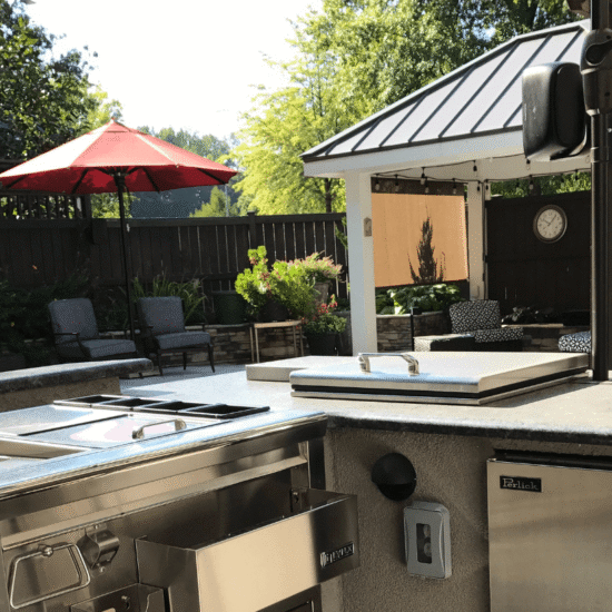 Custom outdoor kitchen and deck with stainless steel appliances, built-in sink, under-counter refrigerator, and integrated storage beneath a shaded structure.