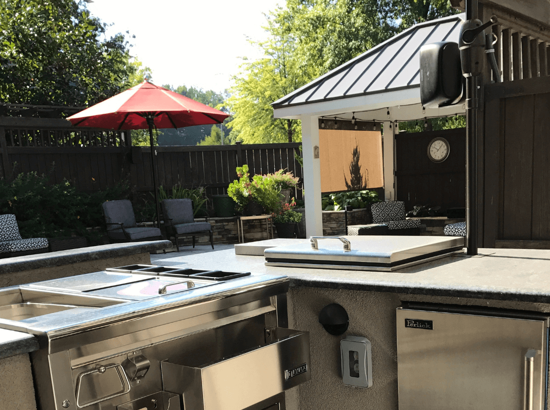 Custom outdoor kitchen and deck with stainless steel appliances, built-in sink, under-counter refrigerator, and integrated storage beneath a shaded structure.